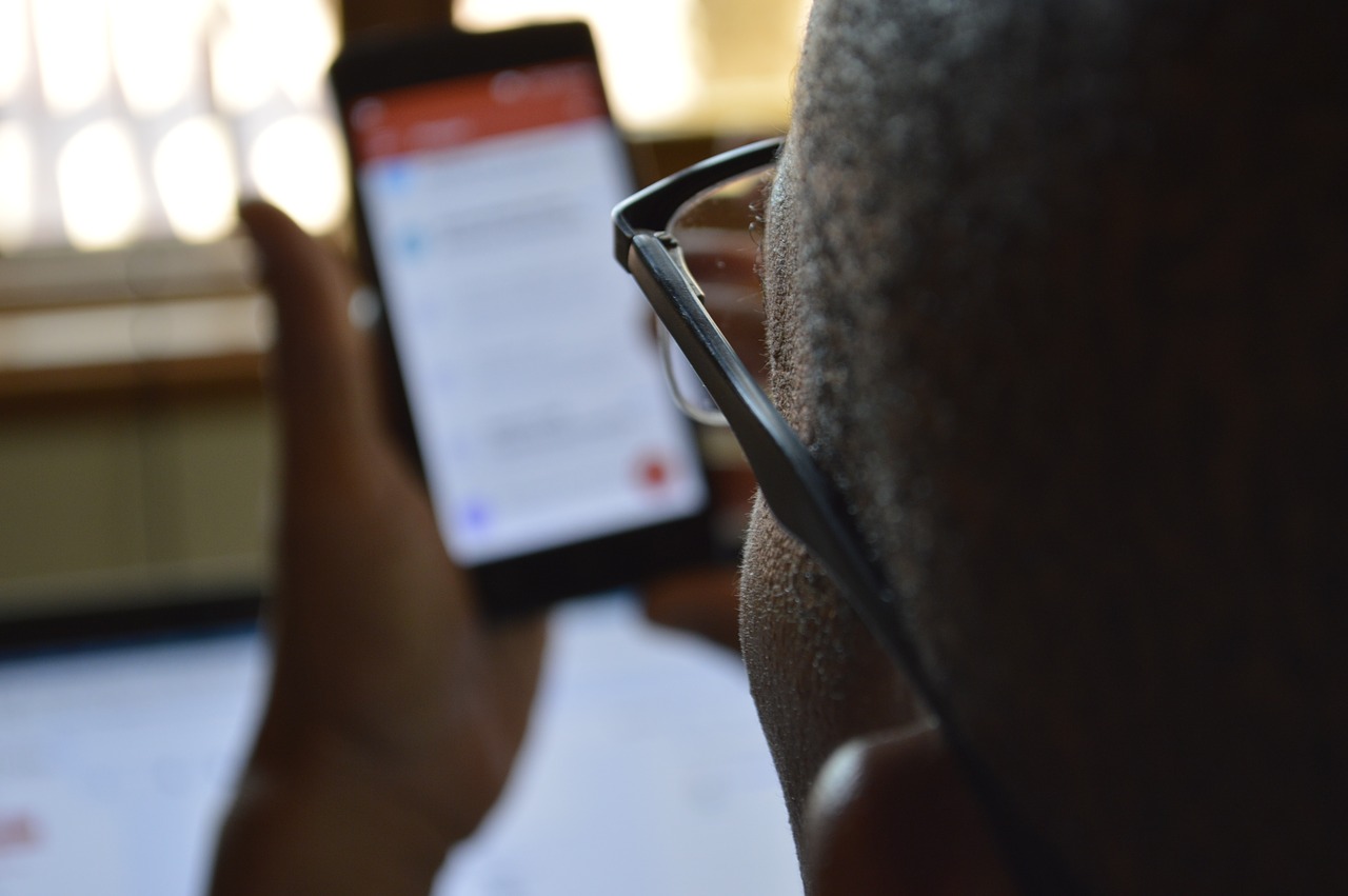 Foto de um homem negro segurando um celular nas mãos. Ele é negro, usa óculos e tem a cabeça raspada.